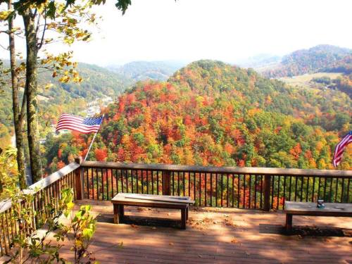 Scenic view from a wooden deck overlooking colorful autumn foliage and rolling hills, with American flags waving.