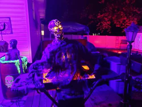 A spooky skeleton with glowing eyes sits on a table, surrounded by Halloween decorations in purple lighting.
