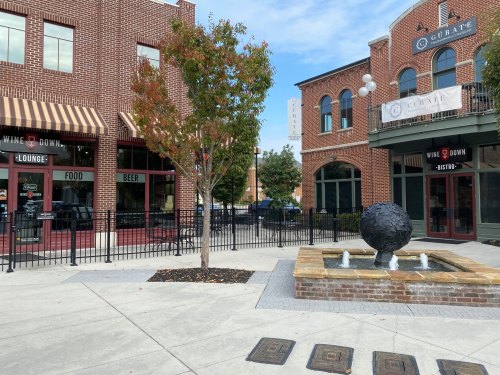 A plaza with a fountain and a tree, surrounded by brick buildings featuring shops and outdoor seating.