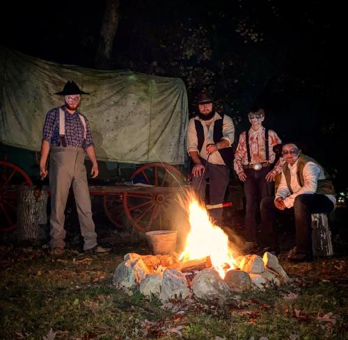 Four people in vintage attire stand around a campfire at night, with a covered wagon in the background.