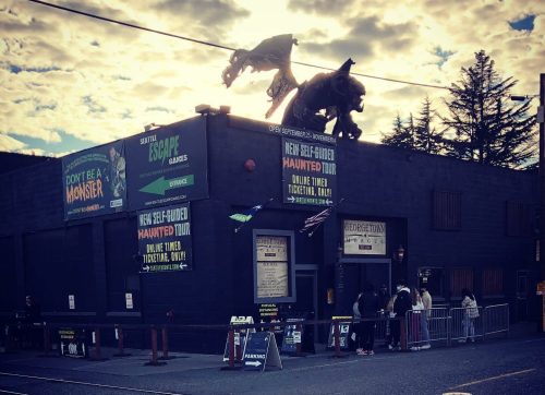 A haunted tour attraction with a dark building, dragon decorations, and a line of people waiting outside under a cloudy sky.