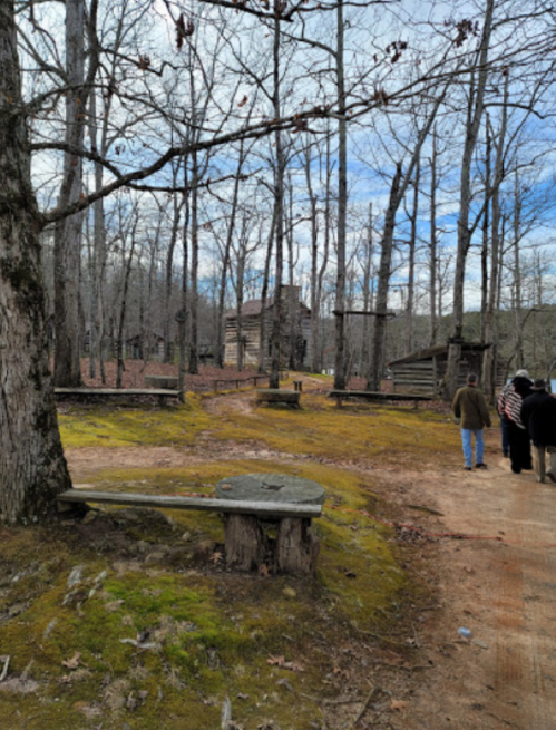 A wooded area with bare trees, a dirt path, and people walking near rustic wooden structures.