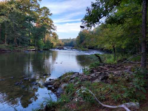 A serene river scene surrounded by trees, with calm water reflecting the sky and rocky banks.