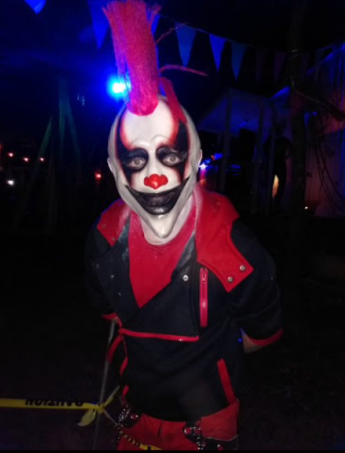 A clown with a mohawk hairstyle, wearing a red and black outfit, stands in a dimly lit, festive environment.