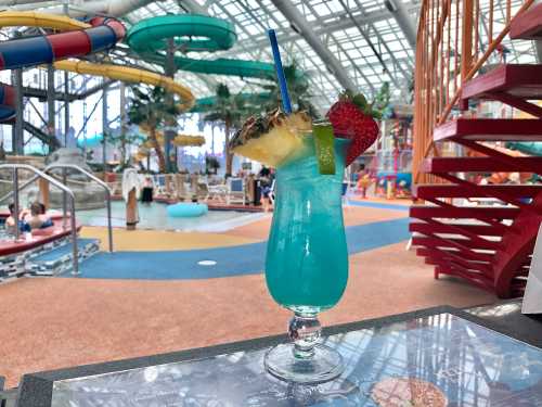 A vibrant blue cocktail garnished with pineapple, strawberry, and lime, set against a colorful water park background.