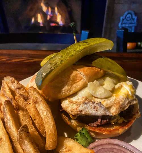 A hearty sandwich with a fried egg, pickles, and fries, served on a plate in front of a cozy fireplace.