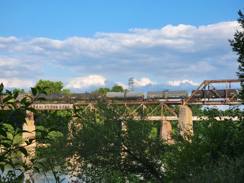 A train crosses a metal bridge over a river, surrounded by greenery and under a blue sky with clouds.