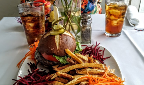 A burger with lettuce, tomato, and a pickle, served with fries and colorful shredded vegetables, alongside drinks.