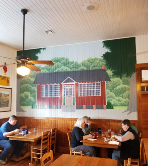 A cozy restaurant interior with a mural of a red cabin and diners enjoying their meals at wooden tables.
