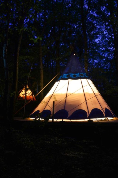 Two illuminated tipis in a forest at night, surrounded by trees and a dark blue sky.