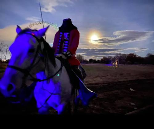 A headless horseman in a red coat rides a horse under a moonlit sky with colorful lights in the background.