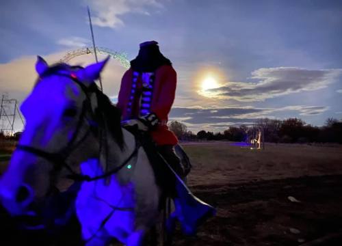 A headless figure in a red coat rides a horse under a moonlit sky with clouds and distant lights.