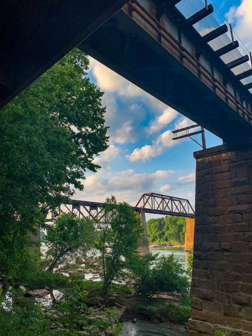 A view of two railway bridges over a river, framed by trees and a blue sky with scattered clouds.