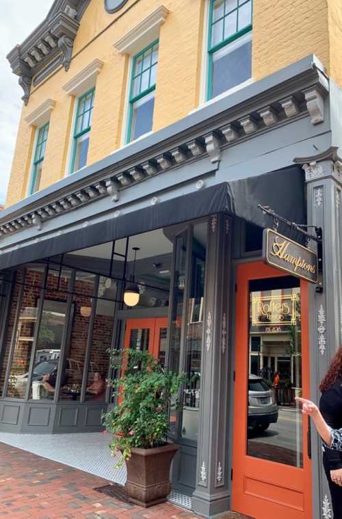 A charming restaurant exterior with yellow walls, large windows, and a black awning, featuring a potted plant at the entrance.