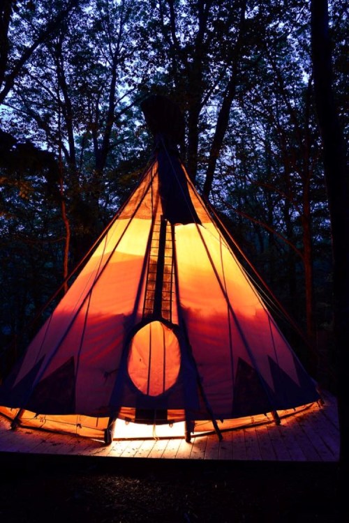 A brightly lit teepee stands in a forest at dusk, surrounded by trees and illuminated by warm orange light.