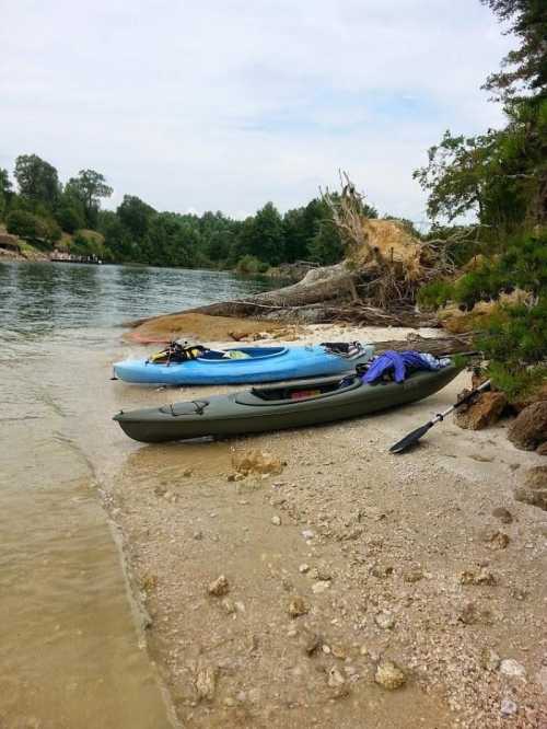 Two kayaks resting on a sandy shore near a calm river, surrounded by trees and a fallen log.