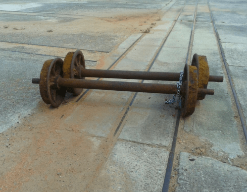 Rusty barbell with weights lying on a concrete surface, partially on metal tracks.