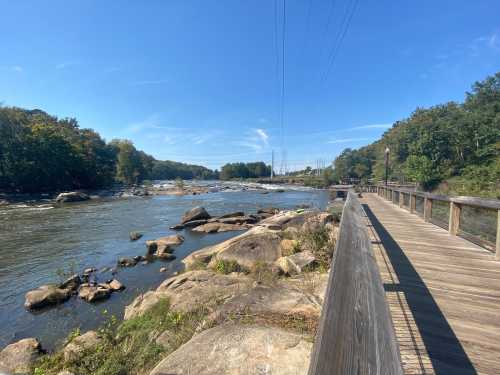 A wooden walkway along a river with rocks, trees, and a clear blue sky in the background.