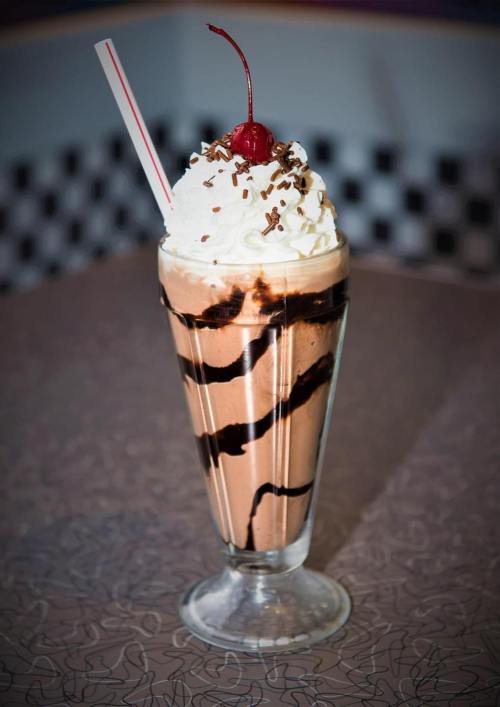 A tall glass of chocolate milkshake topped with whipped cream, chocolate drizzle, and a cherry.