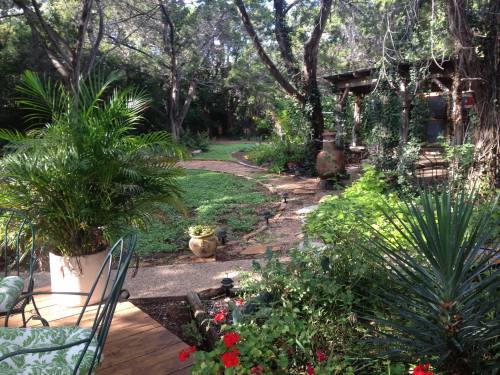 A lush garden path winding through greenery, with potted plants and a cozy seating area in the foreground.