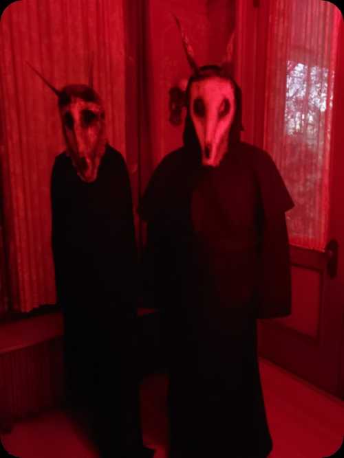 Two figures in dark cloaks wear eerie animal masks, standing in a dimly lit, red-tinted room.