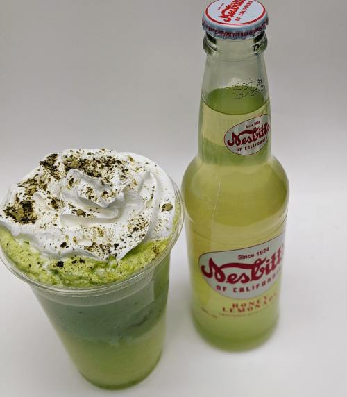 A green layered drink topped with whipped cream and a bottle of Desbitt's soda beside it.
