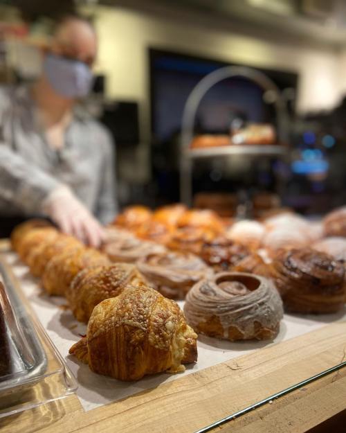 A display of various pastries, including croissants and cinnamon rolls, with a person in the background wearing a mask.