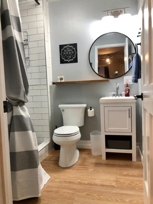 A modern bathroom featuring a shower, toilet, sink with cabinet, and a round mirror against light gray walls.