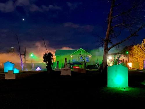 A spooky graveyard at night, illuminated by green lights, with a house in the background and fog rolling in.
