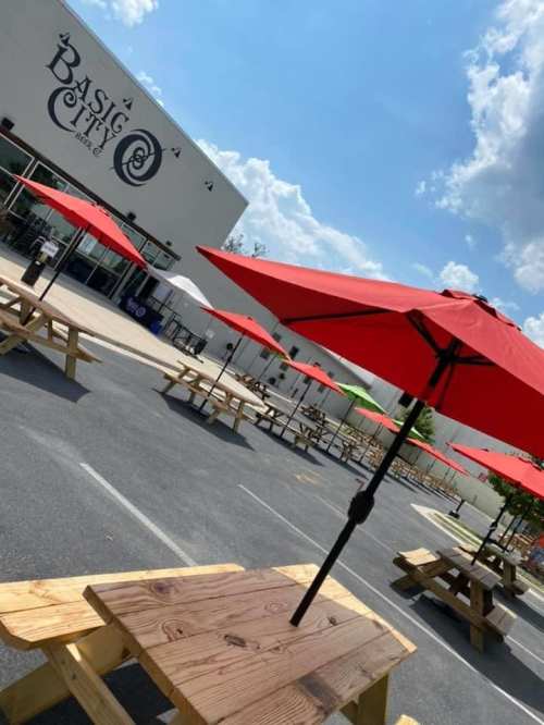 Outdoor seating area with wooden tables and red umbrellas in front of a café under a blue sky with fluffy clouds.