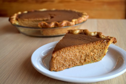A slice of pumpkin pie on a white plate, with the whole pie in a glass dish in the background.