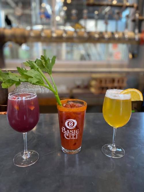 Three colorful cocktails on a bar: a red drink with celery, a purple drink, and a yellow drink with a lemon slice.