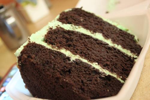 A slice of chocolate cake with green mint frosting, displayed in a takeout container.