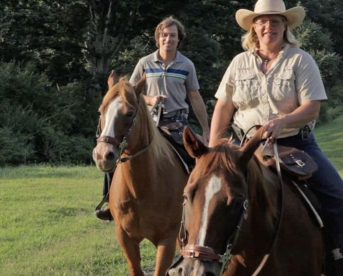 Two riders on horseback in a grassy area, smiling and enjoying a sunny day outdoors.