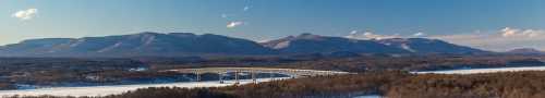 A panoramic view of a bridge spanning a frozen river, surrounded by snow-covered mountains under a clear blue sky.