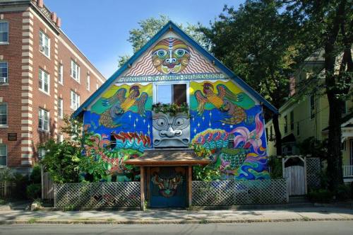 Colorful, mural-covered house with whimsical designs and faces, surrounded by greenery and a decorative fence.