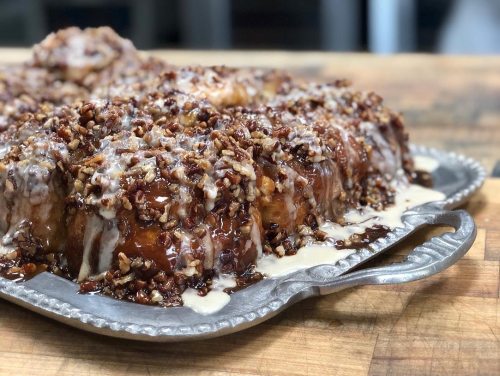 A close-up of a glazed pecan sticky bun on a silver platter, with caramel sauce dripping and nuts sprinkled on top.