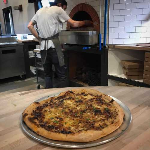 A chef in an apron prepares pizza in a restaurant kitchen, with a freshly baked pizza on a metal tray in the foreground.