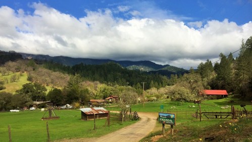 A scenic rural landscape with green fields, a dirt road, and mountains under a partly cloudy sky.