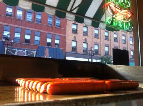 A row of hot dogs on a grill, with a city street visible through a window in the background.