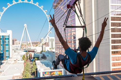 A person zip-lining from a building with a city skyline and a large Ferris wheel in the background, making a peace sign.