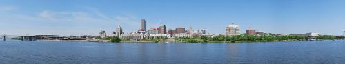 Panoramic view of a city skyline along a river, featuring buildings and greenery under a clear blue sky.