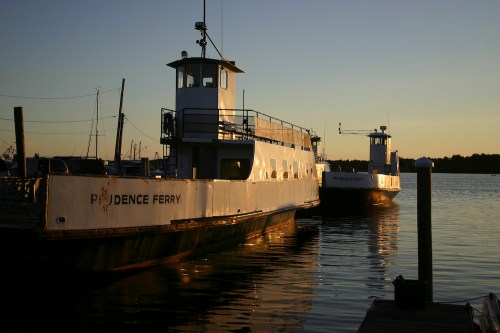 Two ferries docked at sunset, reflecting on calm waters, with silhouettes of boats in the background.