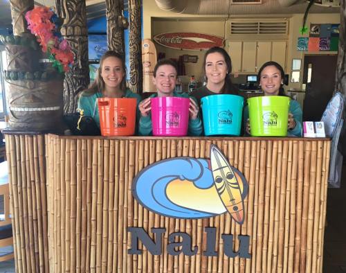Four women stand behind a bamboo bar at Nalu, each holding colorful buckets in front of a surf-themed logo.
