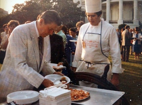 A man in a suit serves himself food from a cart while a chef in a white uniform watches at an outdoor event.