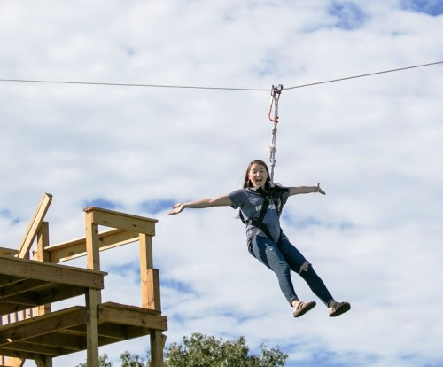 A girl joyfully zip-lining from a wooden platform against a blue sky with clouds.