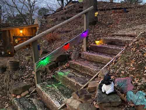 A staircase with colorful lights, surrounded by leaves and stones, leading to a cozy cabin in a wooded area.