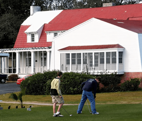 Two men playing golf on a green lawn in front of a large white house with a red roof.