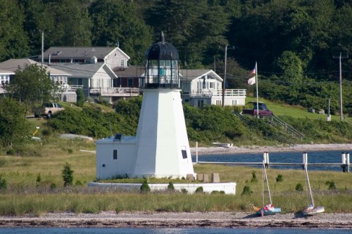 A white lighthouse stands near a grassy shore, with houses and trees in the background and boats on the water.