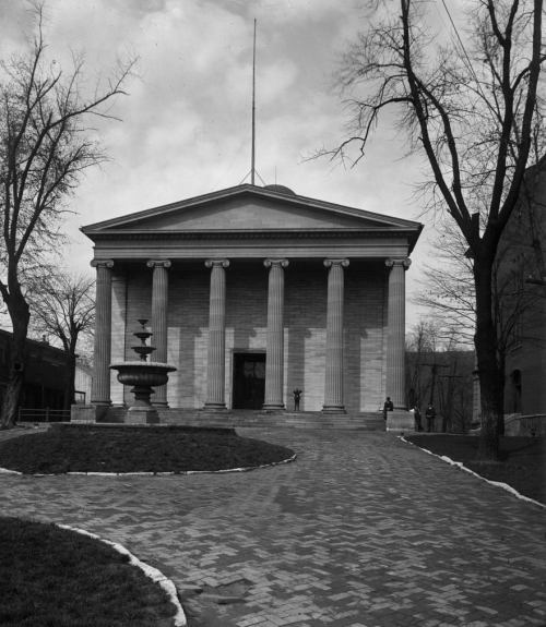 A historic building with columns, a fountain in front, and trees lining the pathway, under a cloudy sky.